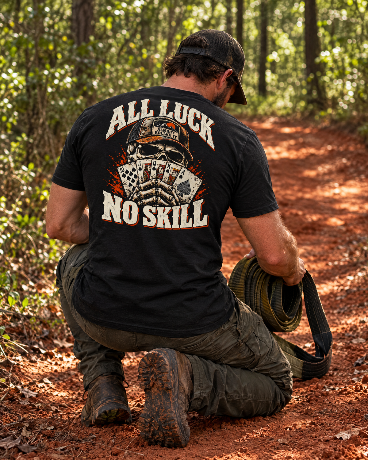 Man kneeling with a recovery strap on a red-clay trail in the All Luck No Skill tee