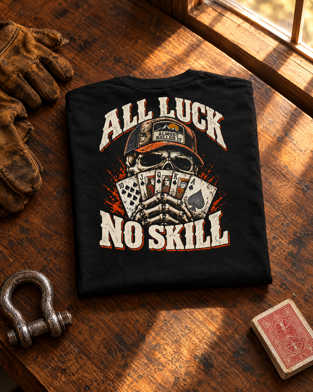 All Luck No Skill tee folded on a walnut workbench next to leather gloves, a shackle and a deck of cards