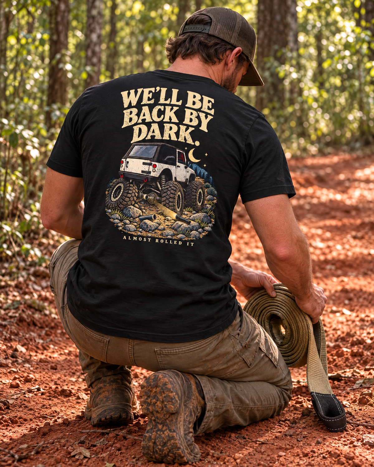 Man kneeling on a red-clay trail coiling a recovery strap, wearing the vintage black Back By Dark tee