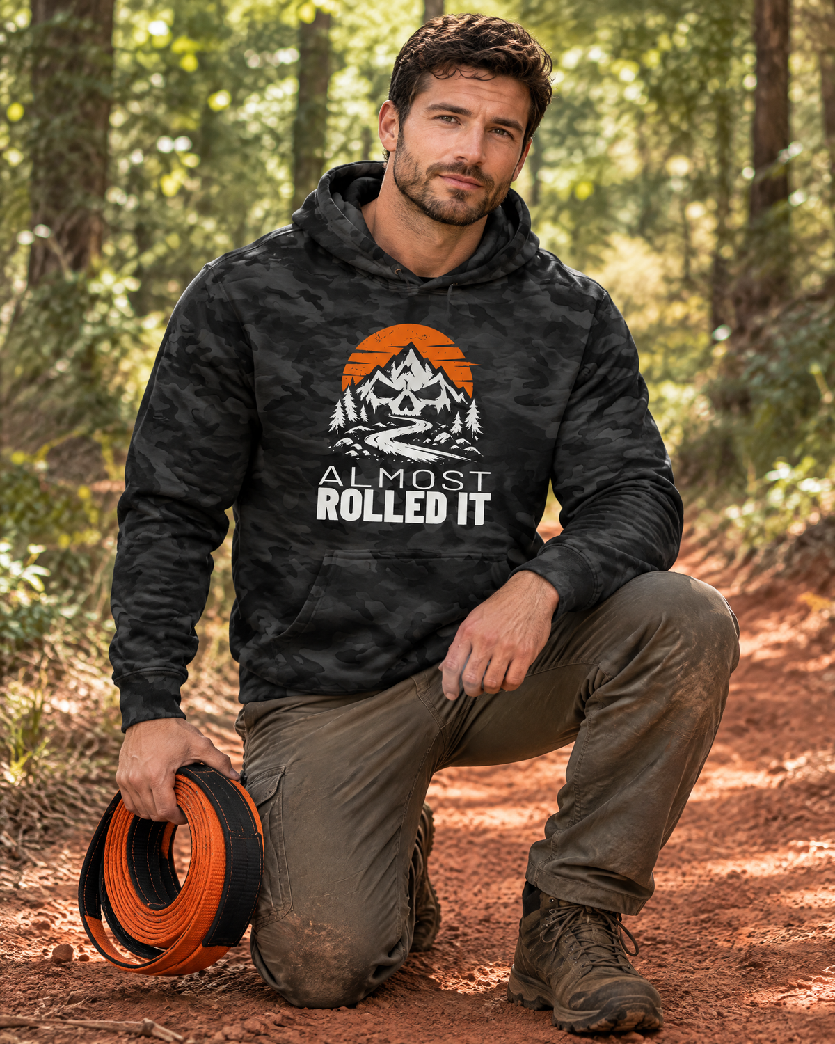 Man in the Almost Rolled It Camo Hoodie kneeling on a red-clay trail with a recovery strap
