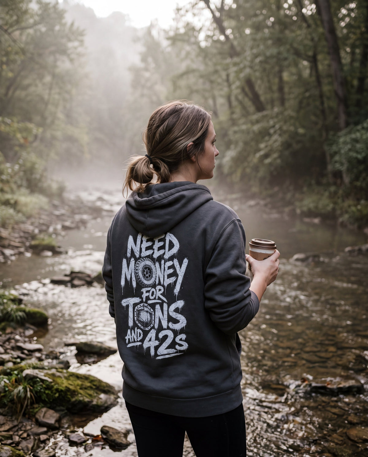 Woman in the asphalt Tons & 42s hoodie at a misty creek, back print showing