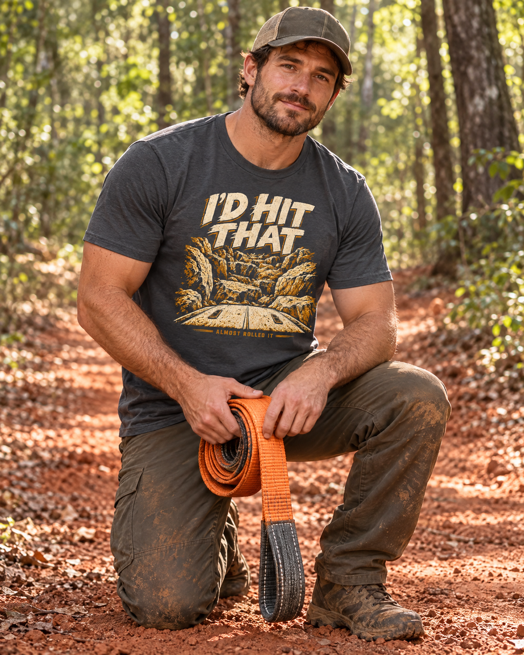 Man kneeling on a red-clay trail holding a recovery strap, wearing the dark grey I'd Hit That tee