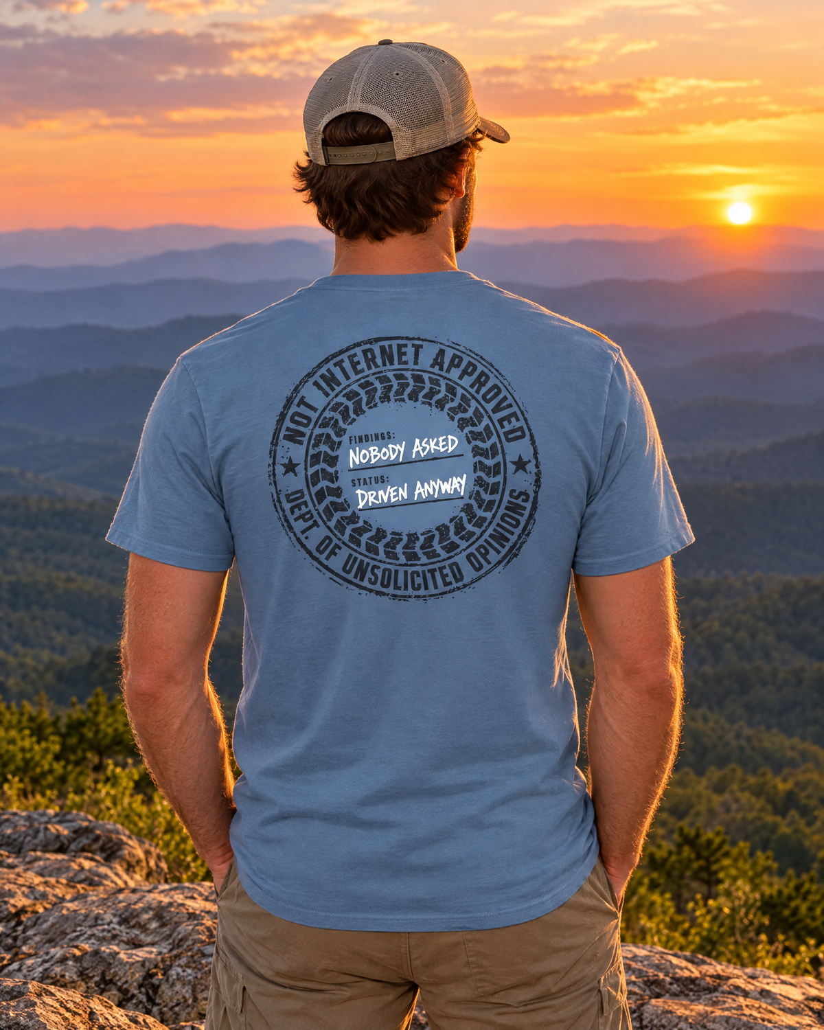 Man wearing the slate Nobody Asked tee on a mountain ledge at sunset, back stamp showing