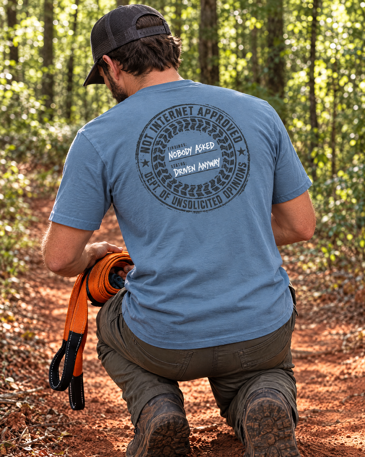 Man coiling a recovery strap on a red-clay trail in the slate Nobody Asked tee