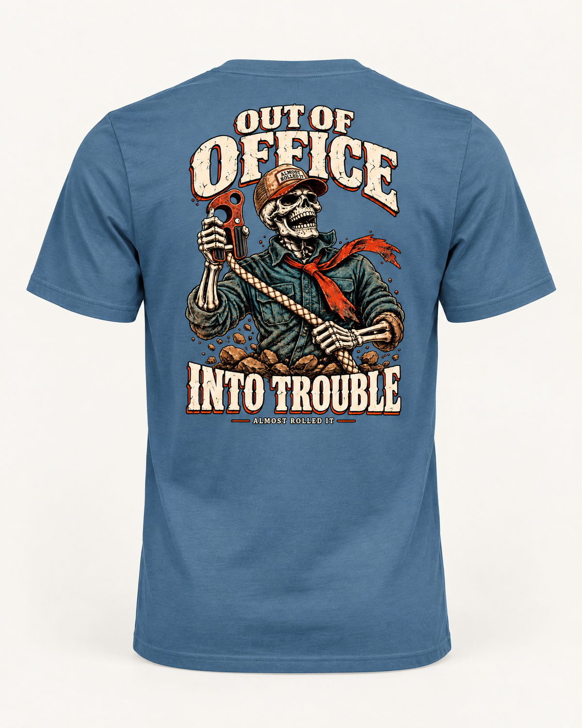 Out of Office tee in steel blue, back view: OUT OF OFFICE, INTO TROUBLE in cream lettering around a laughing skeleton in a trucker cap and red bandana holding a red hook on a rope