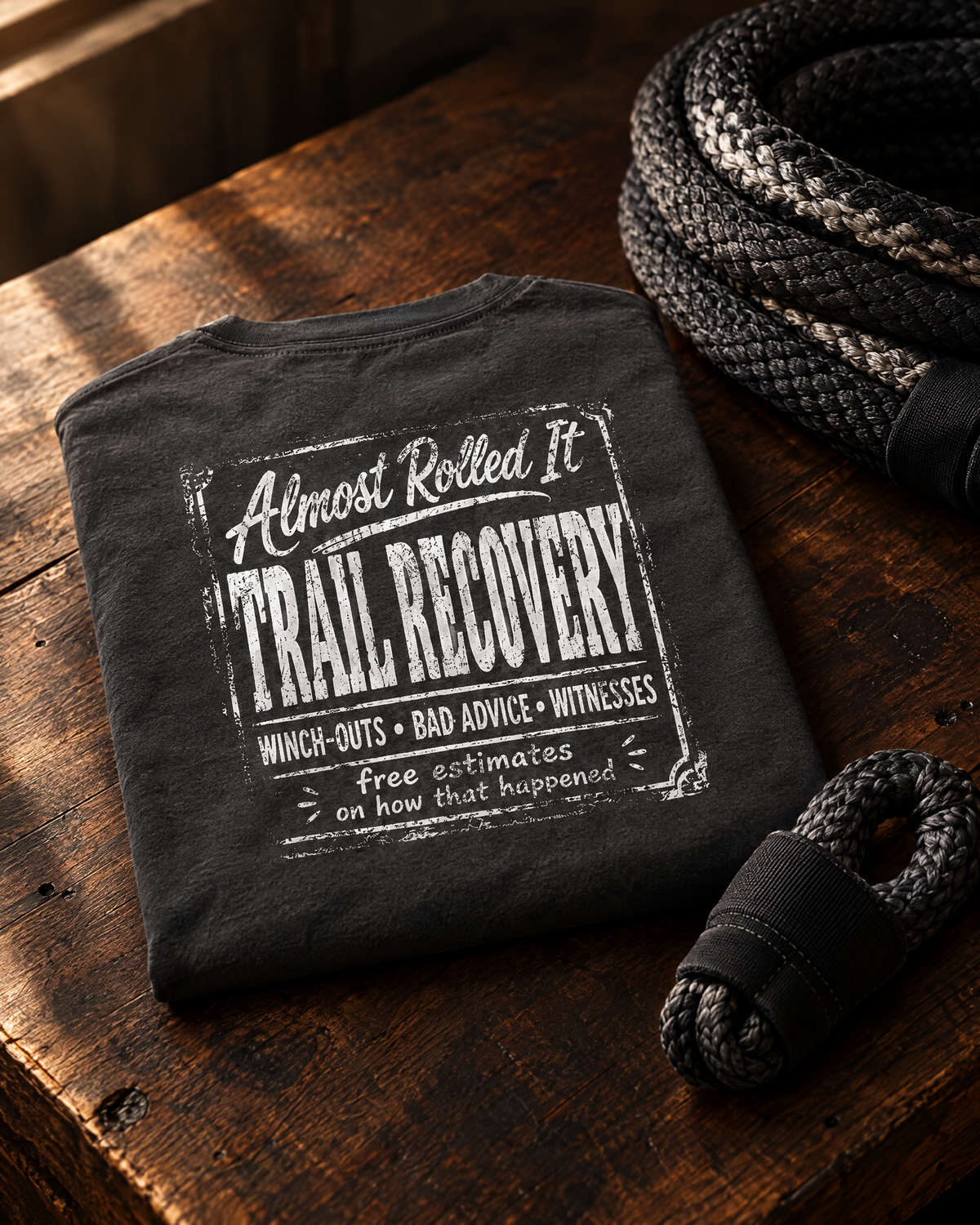 Trail Recovery Badge tee folded on a walnut workbench next to a kinetic recovery rope and soft shackle