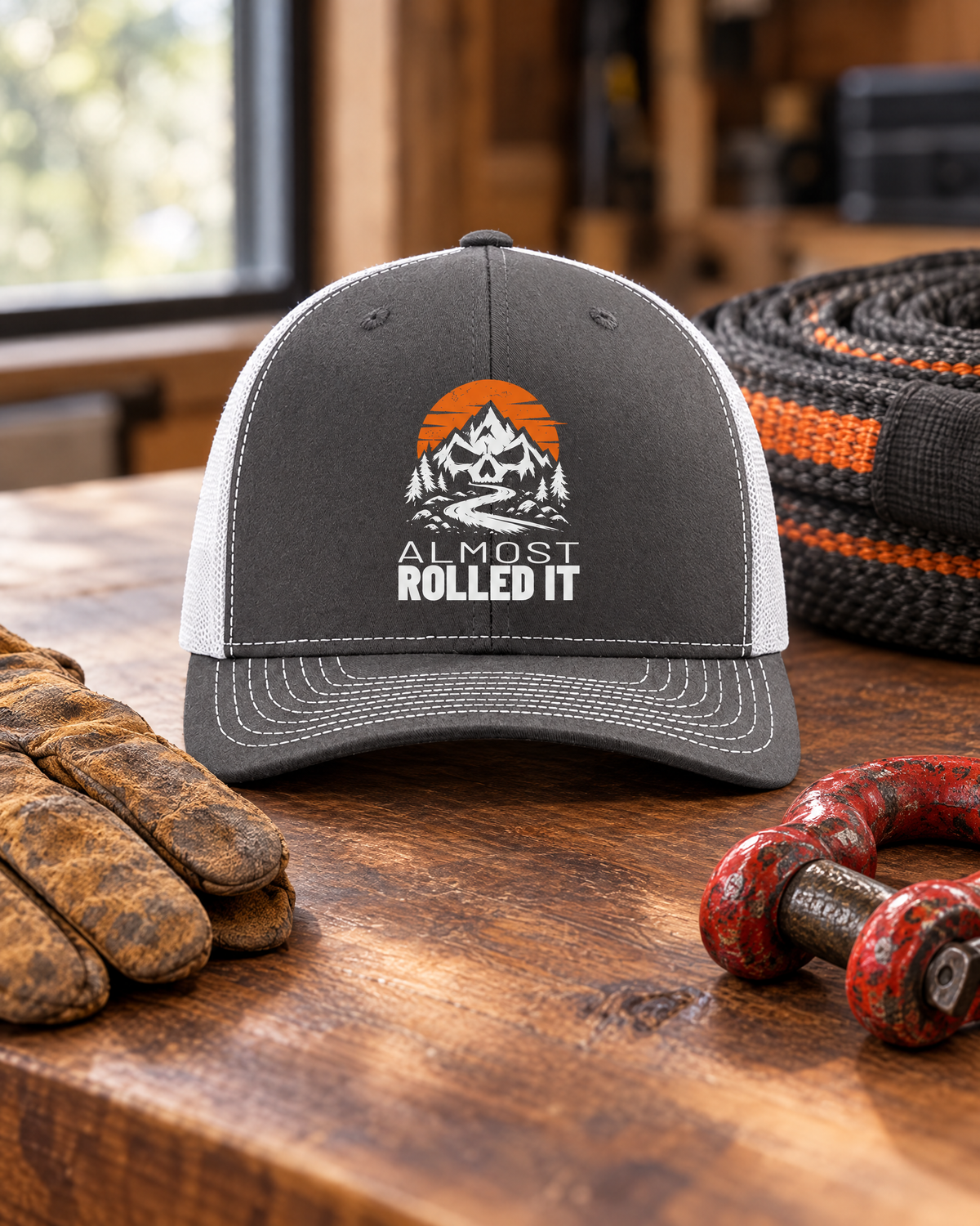 Charcoal and white Almost Rolled It trucker hat on a walnut workbench next to gloves, a shackle and a recovery strap