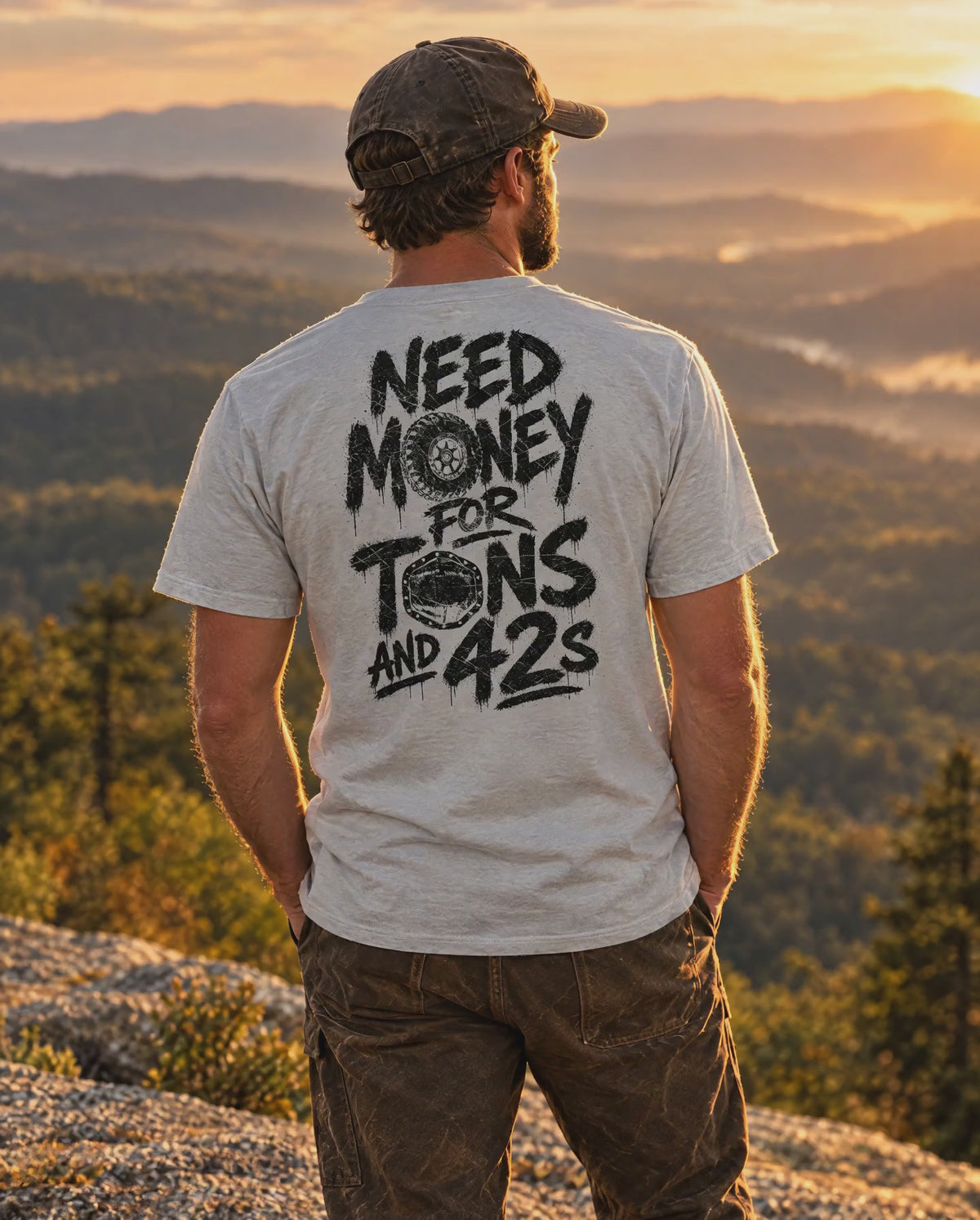 Man in the silver Tons & 42s tee on a mountain ledge at sunset, mist moving in the valley