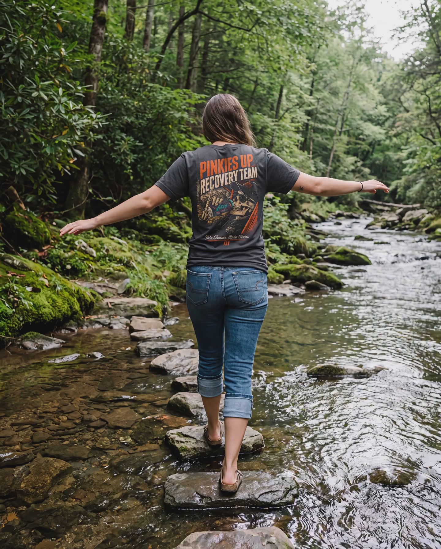 Woman in the Pinkies Up Recovery Team tee balancing across creek stones